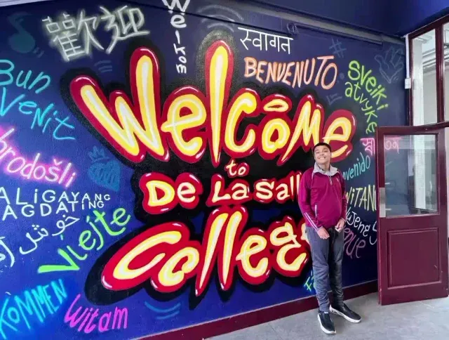 A De La Salle student beside a bright multilingual “Welcome to De La Salle College” mural